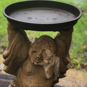May include: A stone-colored statue of a smiling figure with arms raised overhead, supporting a dark brown, oval-shaped plastic tray. The tray features a series of small drainage holes across its base, designed for holding items securely above the figure.