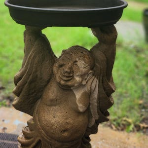 May include: A brown stone statue of a smiling figure with arms raised above the head to support a dark, circular, shallow basin. The figure has a rounded belly and is wearing draped robes. The statue is textured and stands on a circular base.