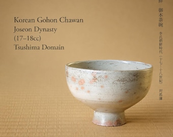May include: A Korean Gohon Chawan tea bowl from the Joseon Dynasty, Tsushima Domain, with a 17-18cc capacity. The ceramic bowl features a light, off-white glaze with subtle orange-toned speckling and a wide, flared rim on a sturdy footed base.