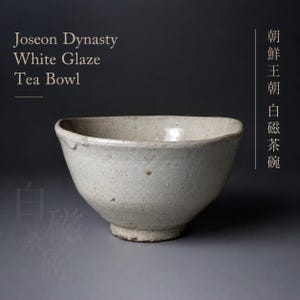 May include: A ceramic tea bowl with a white glaze, featuring a slightly irregular rim and a textured surface with small brown speckles. The text 'Joseon Dynasty White Glaze Tea Bowl' is printed in white on the left side of the image.