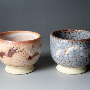 Pode incluir: Duas tigelas de cerâmica feitas à mão. Uma é bege com detalhes em castanho, a outra é azul com uma superfície texturizada. Ambas têm uma base clara e uma borda castanho-avermelhada. As tigelas são provavelmente para servir ou expor.