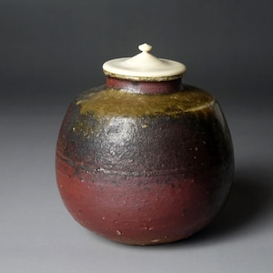 May include: A handcrafted ceramic jar with a lid. The jar has a rounded shape with a dark red base, transitioning to black and olive green near the top. The lid is off-white with a small knob. This decorative piece is suitable for home decor.