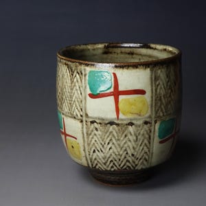 May include: A ceramic tea cup with a textured, off-white background, decorated with colorful geometric shapes. The cup features a brown rim and base, with green, red, and yellow accents. The design includes a herringbone pattern.