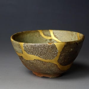 May include: A handcrafted ceramic bowl with kintsugi repair. The bowl has a textured, speckled surface in brown and green, with gold-coloured repair lines. The bowl has a rounded shape and a rustic aesthetic, showcasing the Japanese art of repair.