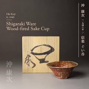 May include: A Shigaraki ware wood-fired sake cup with a textured, mottled brown and reddish surface, positioned in front of a square wooden box featuring black ink calligraphy and a hand-drawn illustration of a cup by artist Oki Koji.
