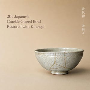 Puede incluir: Un cuenco japonés del siglo XX con acabado de esmalte craquelado gris claro, que presenta líneas de reparación Kintsugi de color dorado visibles en su superficie. El texto 20c Japanese Crackle Glazed Bowl Restored with Kintsugi aparece a la izquierda.