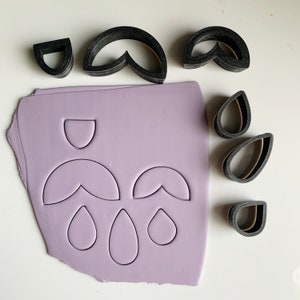 May include: A set of black plastic cookie cutters in various shapes, including a half-moon, a teardrop, and a heart. The cutters are on a purple surface with the shapes cut out of the surface.