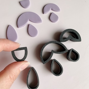 May include: A set of polymer clay cutters in the shape of teardrops and half-circles. The cutters are black and are shown next to purple polymer clay shapes that have been cut out using the cutters.