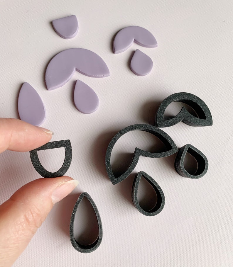 May include: A set of polymer clay cutters in the shape of teardrops and half-circles. The cutters are black and are shown next to purple polymer clay shapes that have been cut out using the cutters.