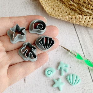 May include: A set of five gray plastic cookie cutters shaped like a starfish, seashell, coral, and two different types of seashells.  Next to the cutters are five small, light blue clay shapes made with the cutters.