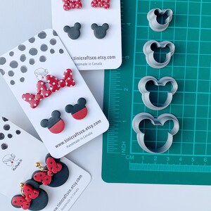 May include: Three sets of polymer clay earrings featuring a red and white polka dot bow and black Mickey Mouse ears. The earrings are displayed on white cards with the Etsy shop name and website address. The image also shows a set of four metal cookie cutters in the shape of Mickey Mouse ears.
