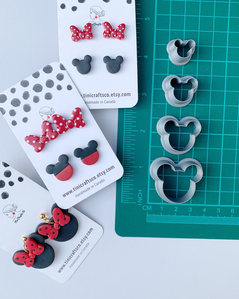 May include: Three sets of polymer clay earrings featuring a red and white polka dot bow and black Mickey Mouse ears. The earrings are displayed on white cards with the Etsy shop name and website address. The image also shows a set of four metal cookie cutters in the shape of Mickey Mouse ears.