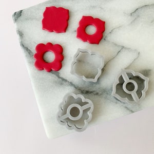 May include: Five cookie cutters on a white marble surface. The cutters are in the shape of flowers with a hole in the center. Two of the cutters are red and three are gray.