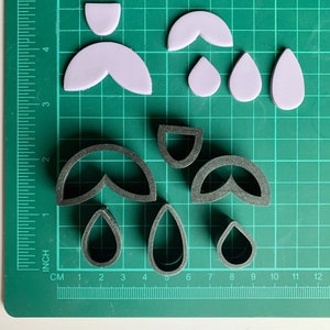 May include: A set of six cookie cutters on a green cutting mat. The cutters are in the shapes of teardrops, half-circles, and a double teardrop shape. The cutters are made of black plastic.