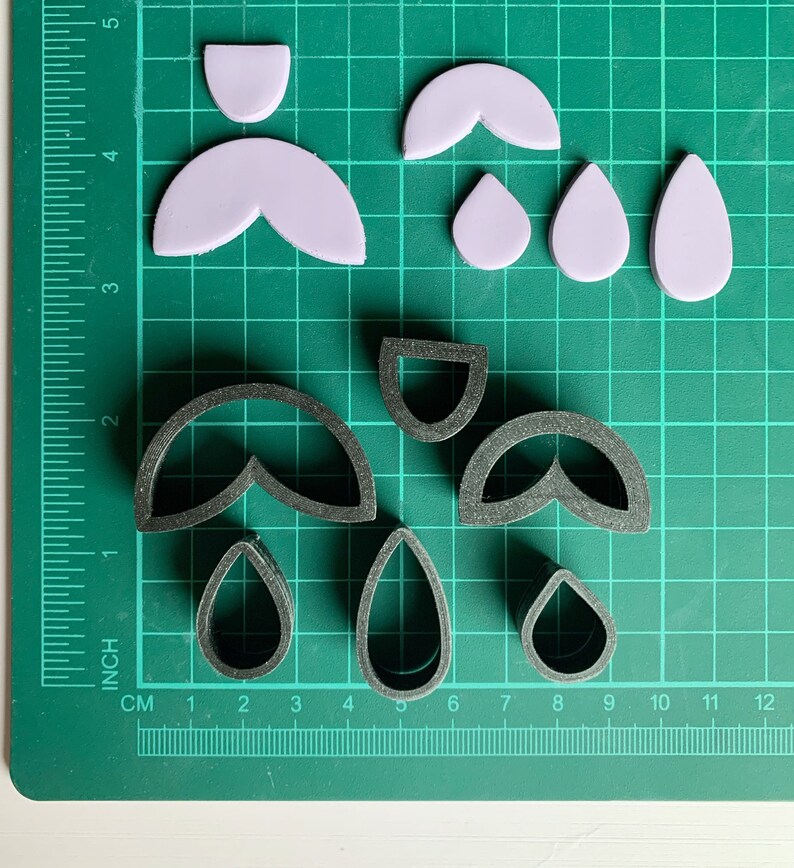 May include: A set of six cookie cutters on a green cutting mat. The cutters are in the shapes of teardrops, half-circles, and a double teardrop shape. The cutters are made of black plastic.