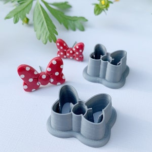 May include: Two grey plastic cookie cutters shaped like bows, one with a red and white polka dot bow inside.