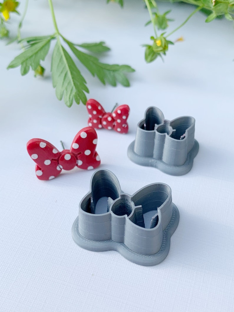 May include: Two grey plastic cookie cutters shaped like bows, one with a red and white polka dot bow inside.