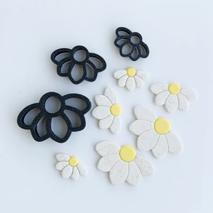 May include: A set of black daisy-shaped cookie cutters and white daisy-shaped polymer clay charms with yellow centres. The charms have a speckled texture.
