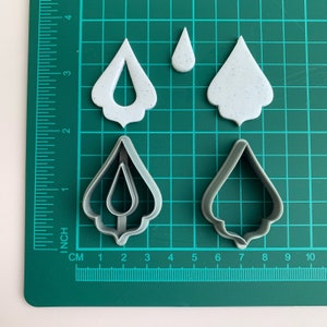 May include: Four cookie cutters in the shape of teardrops. Two are white plastic and two are gray metal. The white cutters have a speckled texture. The metal cutters are smooth. The cutters are on a green cutting mat with inch and centimeter markings.