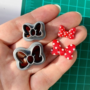 May include: Two grey plastic cookie cutters shaped like bows with a circle in the centre. Two red bows with white polka dots are also shown.