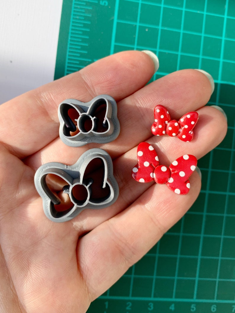 May include: Two grey plastic cookie cutters shaped like bows with a circle in the centre. Two red bows with white polka dots are also shown.