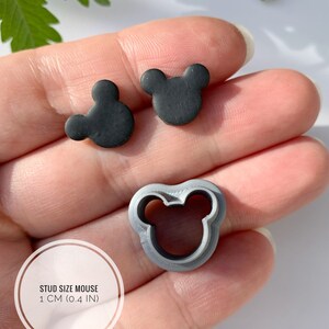 May include: A close-up of a hand holding two black mouse-shaped polymer clay earrings and a grey plastic mouse-shaped cookie cutter. The earrings are 1 cm (0.4 in) in diameter. "STUD SIZE MOUSE 1 CM (0.4 IN)" is written on a white circle.