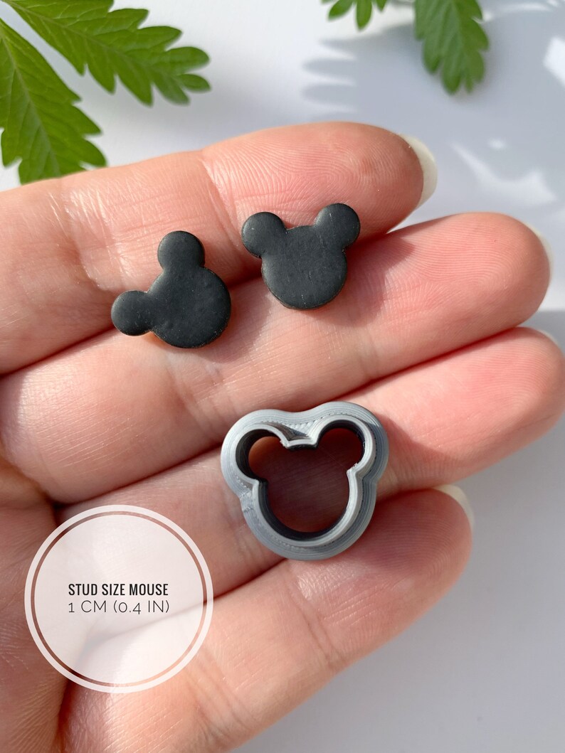 May include: A close-up of a hand holding two black mouse-shaped polymer clay earrings and a grey plastic mouse-shaped cookie cutter. The earrings are 1 cm (0.4 in) in diameter. "STUD SIZE MOUSE 1 CM (0.4 IN)" is written on a white circle.