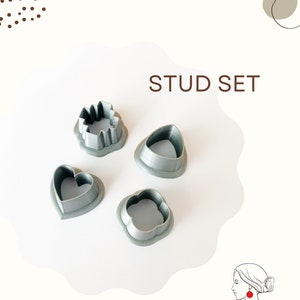May include: A set of four grey plastic stud earring moulds in the shapes of a heart, a flower, a star, and a teardrop. The moulds are arranged on a white surface. The text "STUD SET" is written in brown letters at the top of the image.