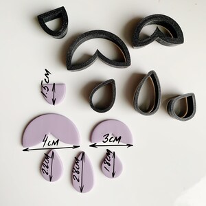 May include: A set of black polymer clay cutters in various shapes, including a teardrop, a half-circle, and a heart. The cutters are arranged on a white surface with purple polymer clay shapes that have been cut out using the cutters. The purple shapes are labeled with their measurements in centimeters.