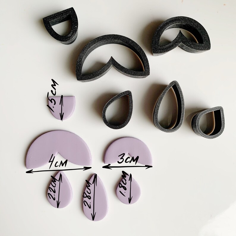 May include: A set of black polymer clay cutters in various shapes, including a teardrop, a half-circle, and a heart. The cutters are arranged on a white surface with purple polymer clay shapes that have been cut out using the cutters. The purple shapes are labeled with their measurements in centimeters.
