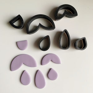 May include: A set of polymer clay cutters and a selection of purple polymer clay shapes. The cutters are black and have various shapes, including teardrops, half circles, and a flower shape. The polymer clay shapes are teardrops, half circles, and a flower shape.