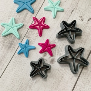 May include: A set of eight cookie cutters in the shape of a starfish. The cutters are in three different sizes and are made of grey plastic. There are also three starfish-shaped cookies made of fondant in blue, pink, and light blue.