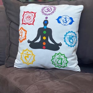 May include: White square pillow featuring a black silhouette of a person in a seated yoga pose. The pillow has colorful chakra symbols and dots in red, orange, yellow, green, blue, and purple. The pillow is on a brown couch.