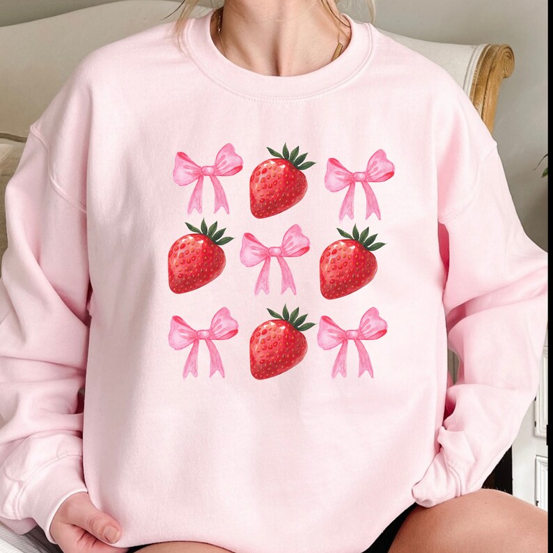 K&ouml;nnte beinhalten: Hellrosa Sweatshirt mit einem Muster aus roten Erdbeeren und rosa Schleifen. Die Erdbeeren haben gr&uuml;ne Bl&auml;tter. Die Schleifen sind in einem helleren Rosa gehalten. Das Design befindet sich auf der Vorderseite des Sweatshirts.