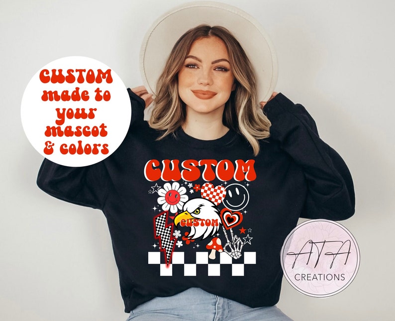 Op de afbeelding: Zwart sweatshirt met een aangepast ontwerp met een adelaar, bloemen, harten en een geblokte vlag. Het woord "CUSTOM" is rood en het ontwerp bevat de tekst "CUSTOM made to your mascot & colors."
