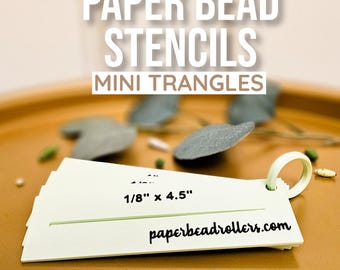May include: A set of pale green paper bead stencils labeled MINI TRANGLES and 1/8" x 4.5" with the website paperbeadrollers.com printed on the surface. The stencils are held together by a pale green ring and feature a horizontal slit for cutting.
