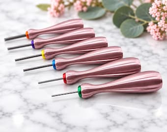 Puede incluir: Un juego de seis punzones de metal con mangos de oro rosa metálico y puntas de acero finas. Cada herramienta presenta un anillo de color distinto en la base del mango, incluyendo acentos en naranja, morado, amarillo, azul, rojo y verde.