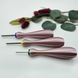May include: Three pink screwdrivers with coloured tips: purple, yellow, and green. The handles have a ribbed design. The tools are arranged on a white surface with greenery in the background.