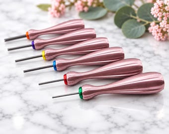 Puede incluir: Un juego de seis punzones de metal con mangos de oro rosa metálico y puntas de acero finas. Cada herramienta presenta un anillo de color distinto en la base del mango, incluyendo acentos en naranja, morado, amarillo, azul, rojo y verde.