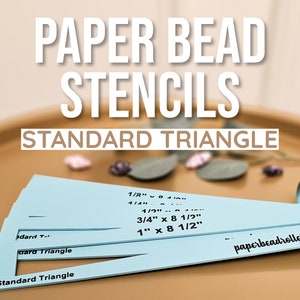 May include: A set of blue paper bead stencils for making triangles. The stencils are labeled with different sizes, including 1/8 inch, 1/2 inch, 3/4 inch x 8 1/2 inch, and 1 inch x 8 1/2 inch. The stencils are on a brown surface with the text "paperbeadrollers.com" on the bottom right corner.