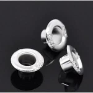 May include: Three silver eyelets with a decorative, textured rim and a shiny, smooth interior. The eyelets are set against a dark, reflective surface, highlighting their metallic sheen. These eyelets are likely used for crafting or garment construction.
