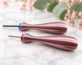 Puede incluir: Dos herramientas de metal con mangos ergonómicos en tono oro rosa con ranuras verticales. La herramienta superior tiene un collar azul cerca de la punta metálica fina y recta, mientras que la inferior tiene un collar rojo cerca de la punta.