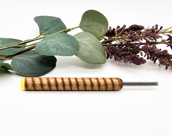 Puede incluir: Una herramienta de madera con un mango de ranuras en espiral y una tapa final de color amarillo brillante. Una varilla de metal delgada de color plateado se extiende desde el extremo opuesto del mango sobre un fondo blanco liso.