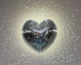 May include: A heart-shaped disco ball with a mirrored surface, reflecting light. The ball is centered on a white wall, surrounded by a pattern of square light reflections. The overall effect is a sparkling, radiant display.