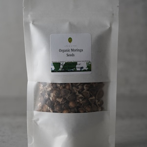 Puede incluir: Una bolsa blanca resellable con una etiqueta que dice "Organic Moringa Seeds" y enumera los beneficios nutricionales de las semillas de moringa. La bolsa contiene semillas de moringa marrones.
