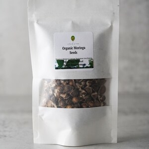 Puede incluir: Una bolsa blanca resellable con una etiqueta que dice "Organic Moringa Seeds" y enumera los beneficios nutricionales de las semillas de moringa. La bolsa contiene semillas de moringa marrones.