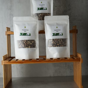 Puede incluir: Tres bolsas blancas de semillas de moringa org&aacute;nica en un estante de madera. Las bolsas tienen una ventana transparente que muestra las semillas en el interior.