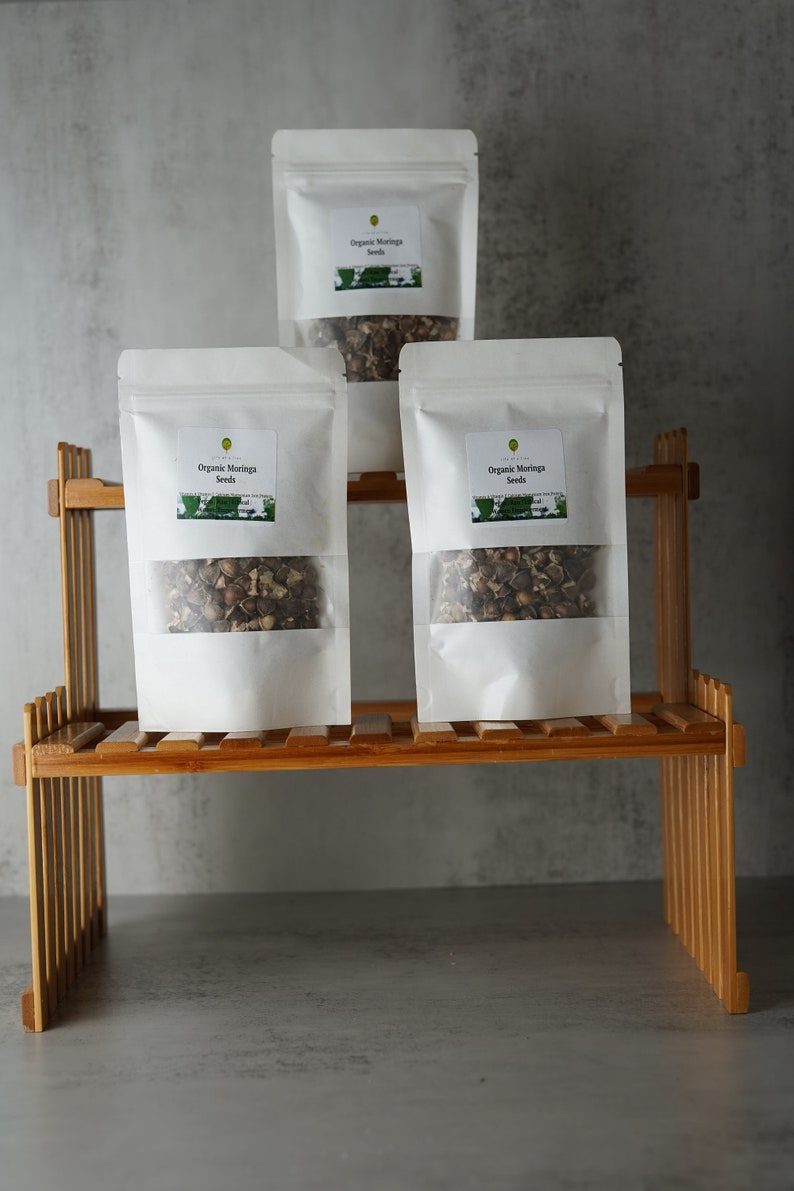 Puede incluir: Tres bolsas blancas de semillas de moringa org&aacute;nica en un estante de madera. Las bolsas tienen una ventana transparente que muestra las semillas en el interior.