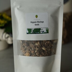 Puede incluir: Una bolsa blanca resellable con una etiqueta que dice "Life of a Tree Organic Moringa Seeds" y enumera los beneficios nutricionales de las semillas de moringa. La bolsa contiene semillas de moringa marrones.