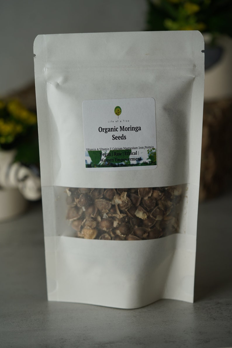 Puede incluir: Una bolsa blanca resellable con una etiqueta que dice "Life of a Tree Organic Moringa Seeds" y enumera los beneficios nutricionales de las semillas de moringa. La bolsa contiene semillas de moringa marrones.